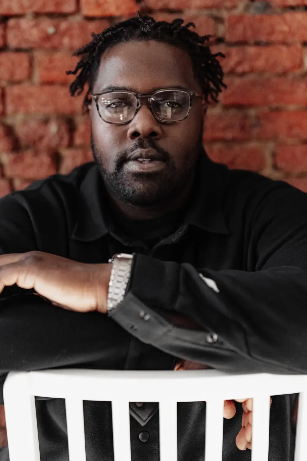 A man with glasses and a black shirt sits behind a white chair, resting his arms on the chair back, with a red brick wall in the background.