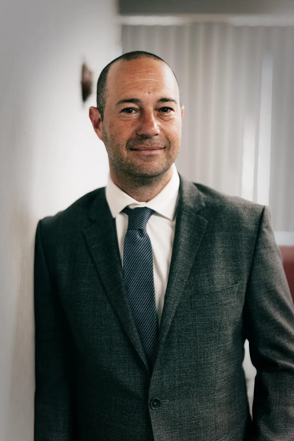 A man in a gray suit, white shirt, and blue tie stands indoors, leaning against a white wall with vertical blinds in the background.